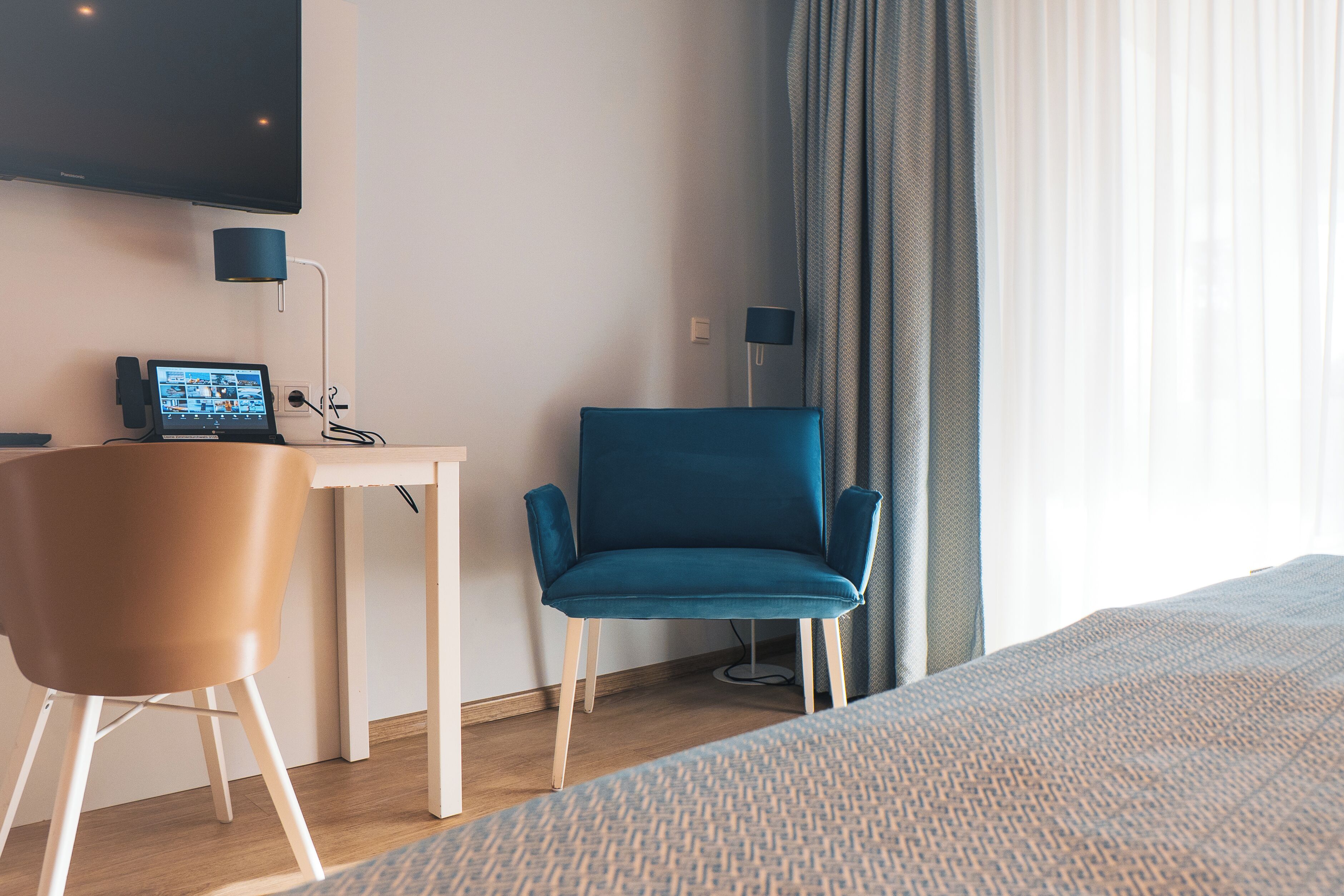 A hotel room with a bed, a desk with a computer, a blue chair, and a TV mounted on the wall.