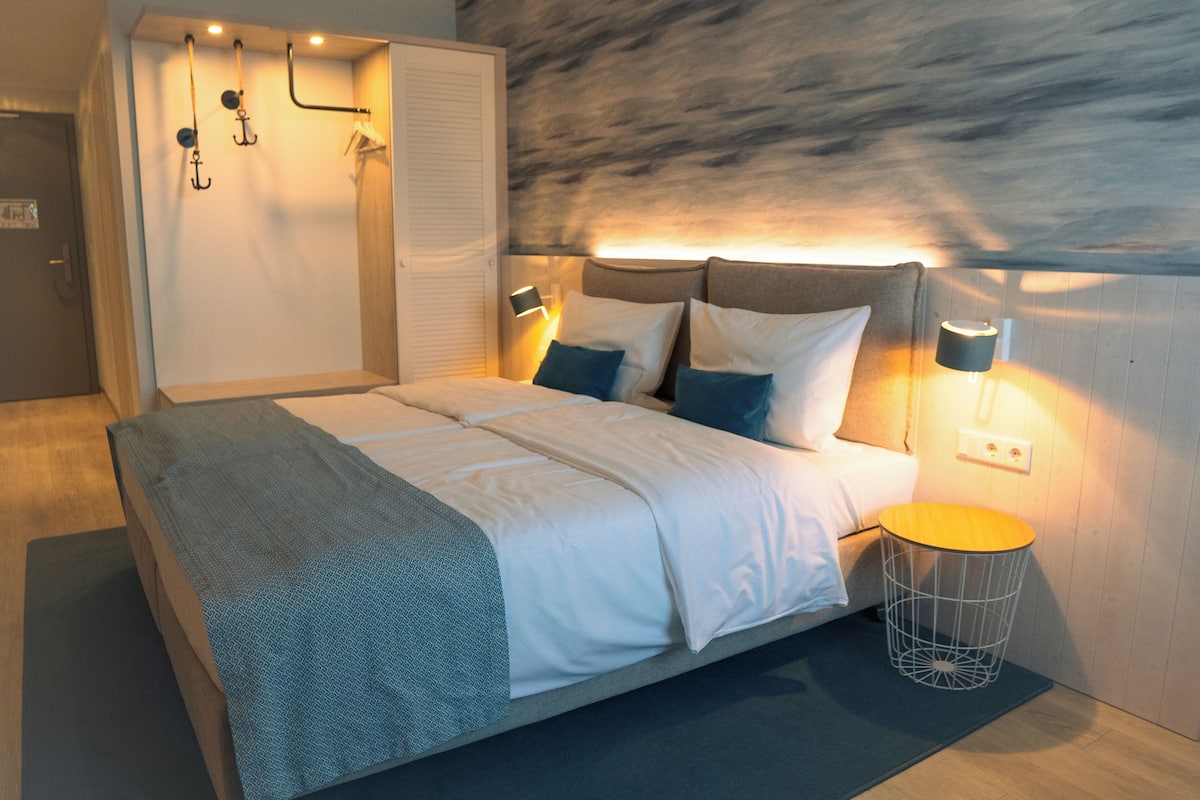 A modern bedroom with a bed, bedside lamps, a hanging light fixture, and a wooden floor.
