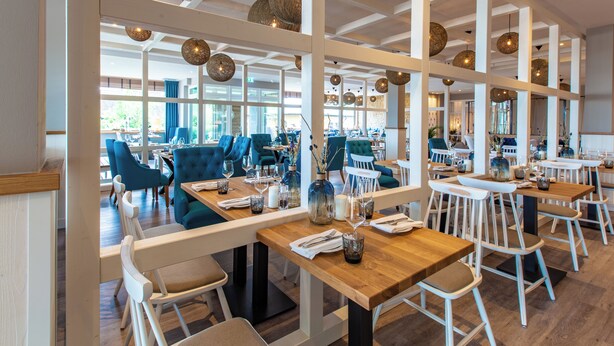 Un restaurante moderno con mesas de madera y sillas blancas, ventanas amplias y lámparas esféricas colgantes.
