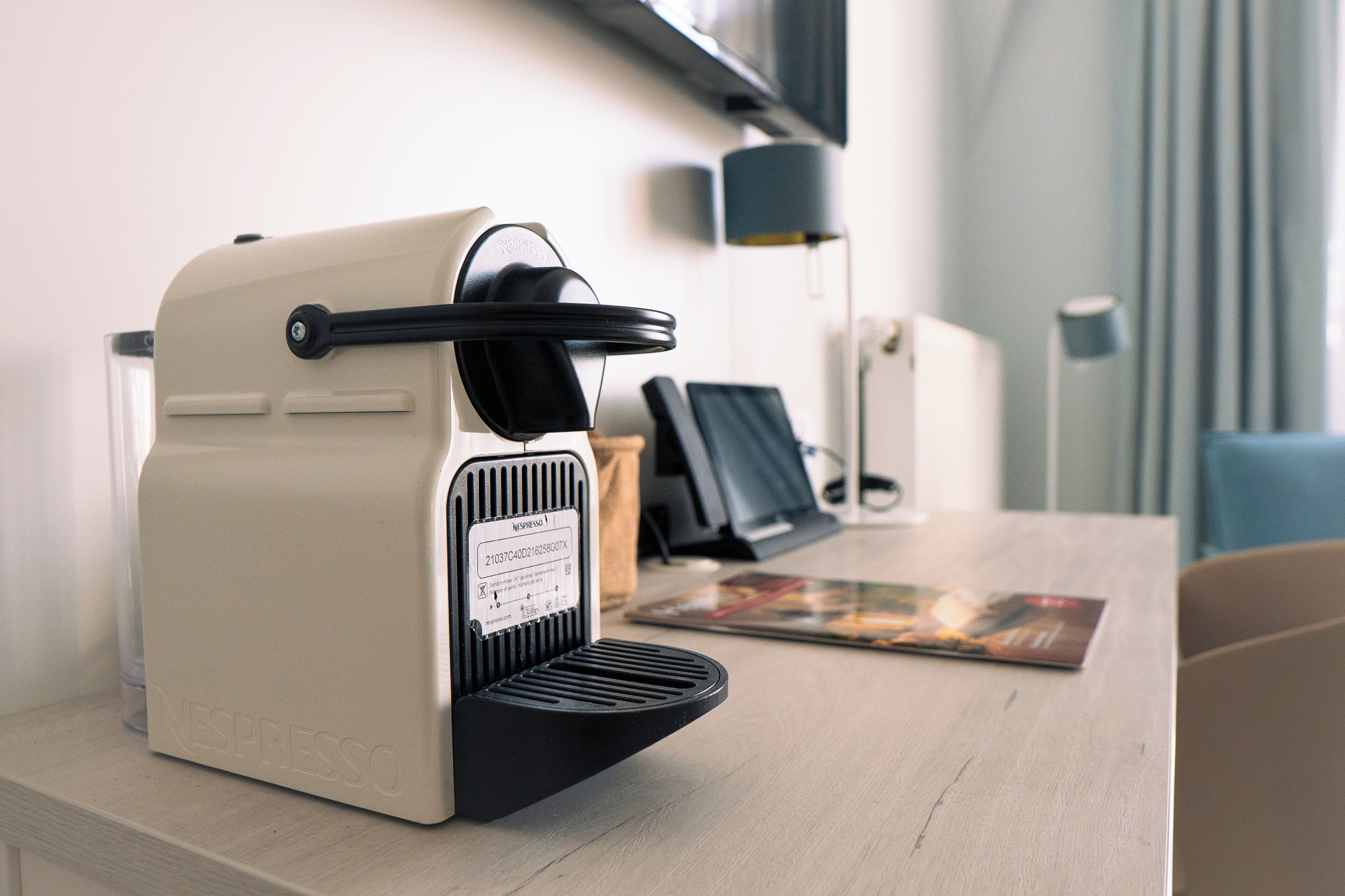 Een Nespresso koffiemachine op een houten tafel, met een tijdschrift en een lamp op de achtergrond.