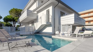 A modern two-story building with a swimming pool, outdoor seating, and a white fence. - Modern Apartment With Shared Pool (San Michele al Tagliamento)