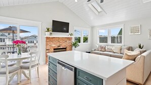 Una cocina espaciosa con una isla central, una zona de comedor con sillas blancas, una chimenea de ladrillo empotrada y un amplio ventanal con vistas a una zona residencial.