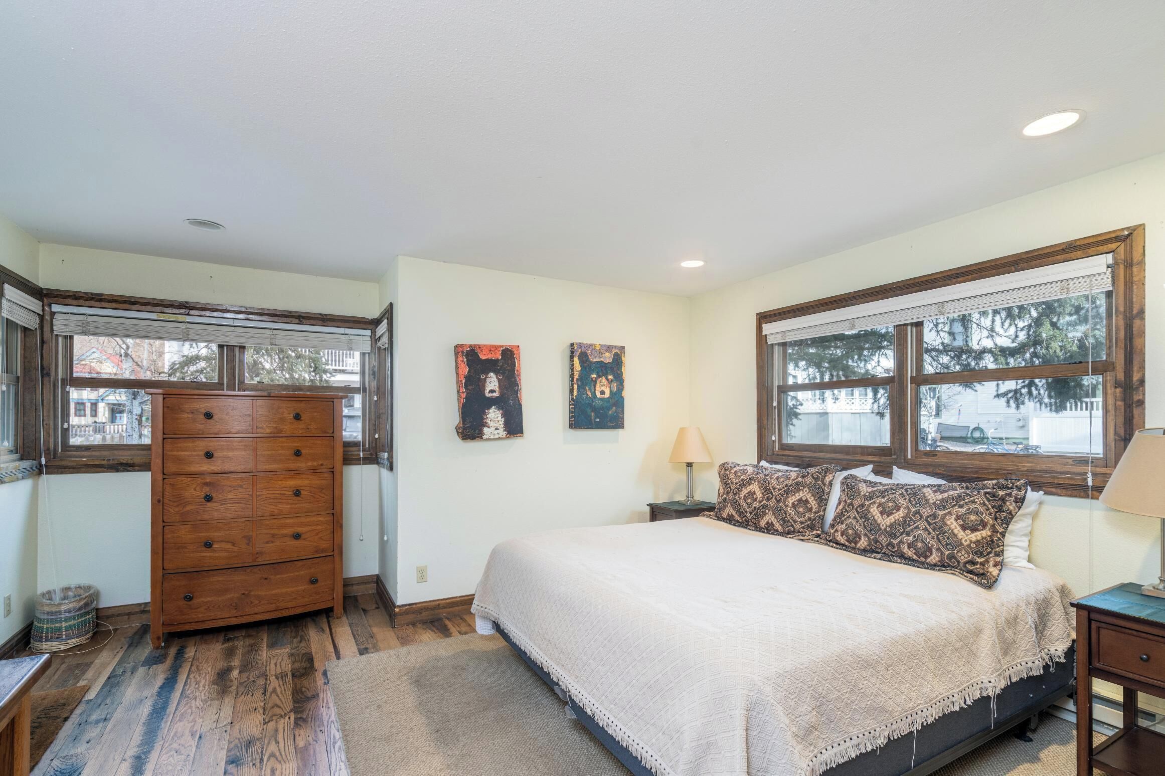 Une chambre avec un grand lit, une commode en bois, deux fenêtres et une vue sur l’extérieur.