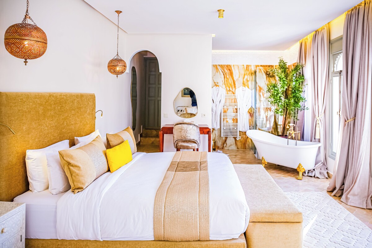 A spacious bedroom with a large bed, a sitting area, a bathtub, and a view of a cityscape.