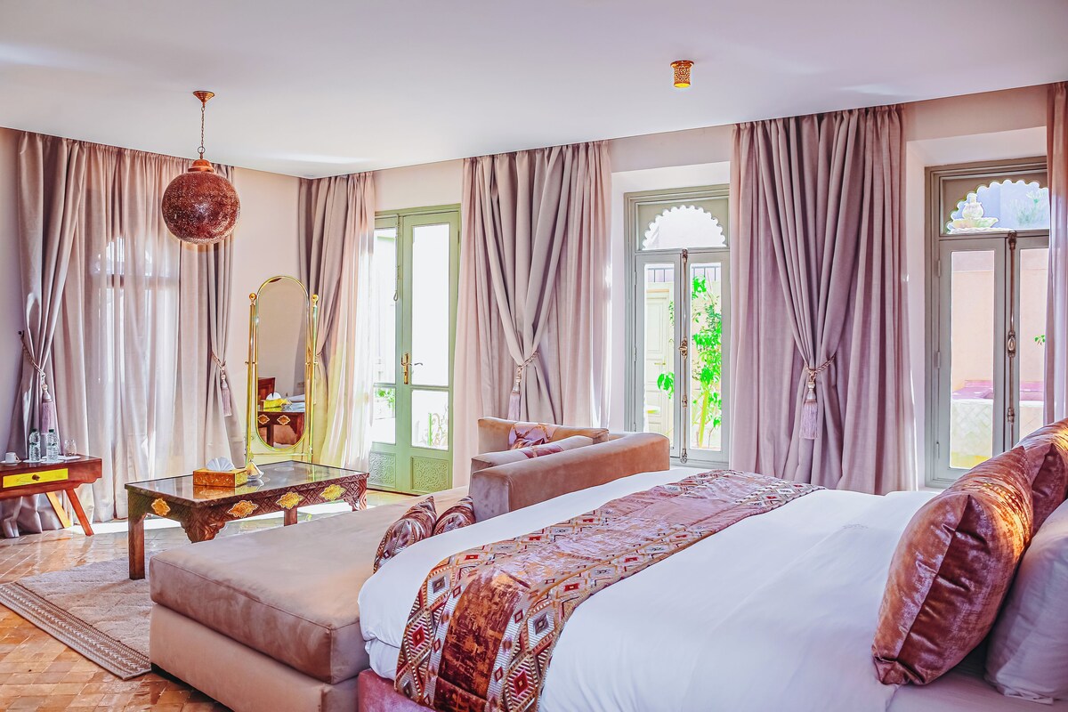 A bedroom with a large bed, a sitting area, a vanity with a mirror, and a view of greenery outside.