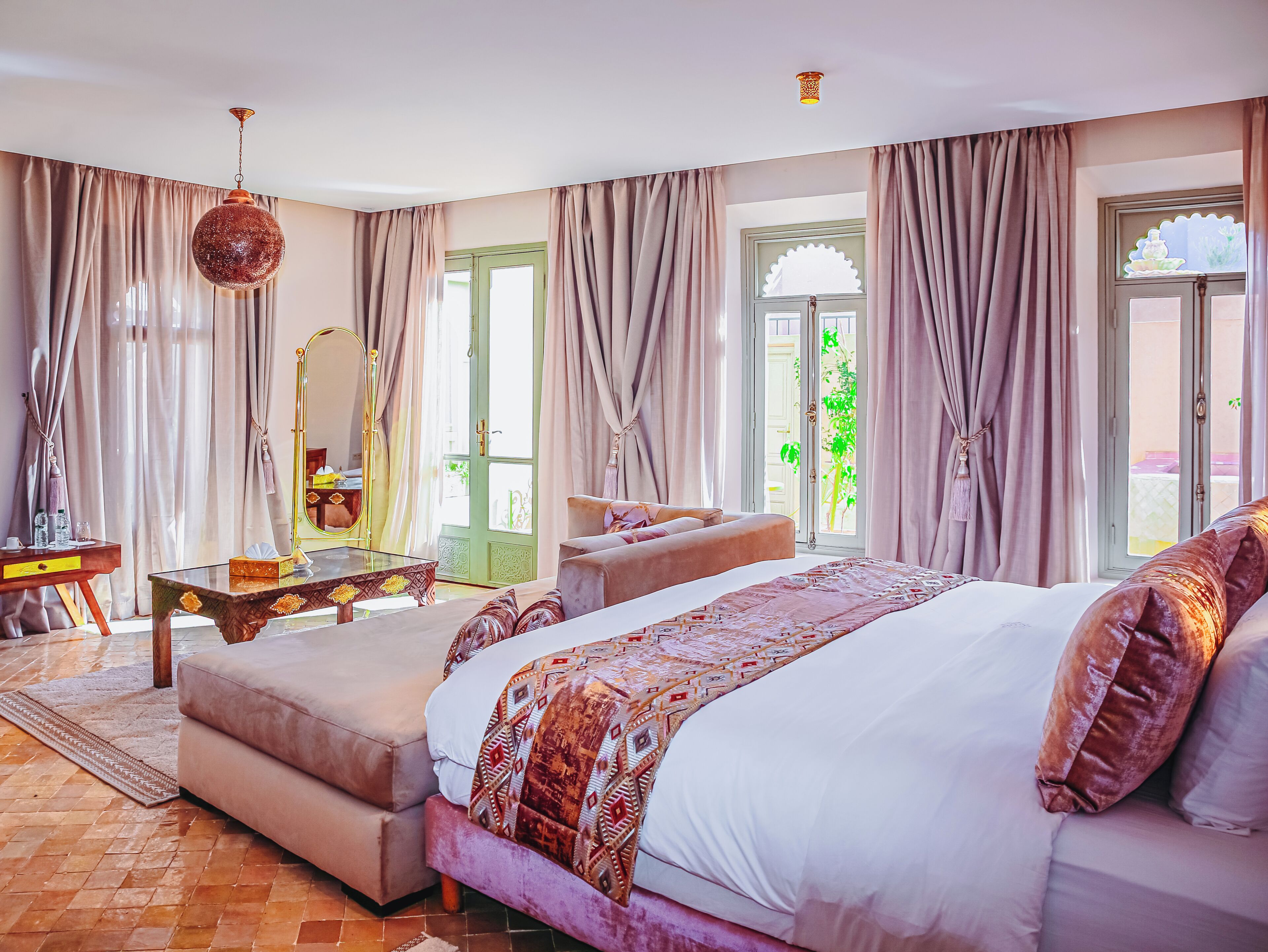 A bedroom with a large bed, a sitting area, a vanity with a mirror, and a view of greenery outside.