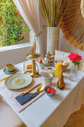 Una mesa de desayuno dispuesta con platos, cubiertos, un calentador de agua para té, un vaso de jugo de naranja y una flor en un florero.