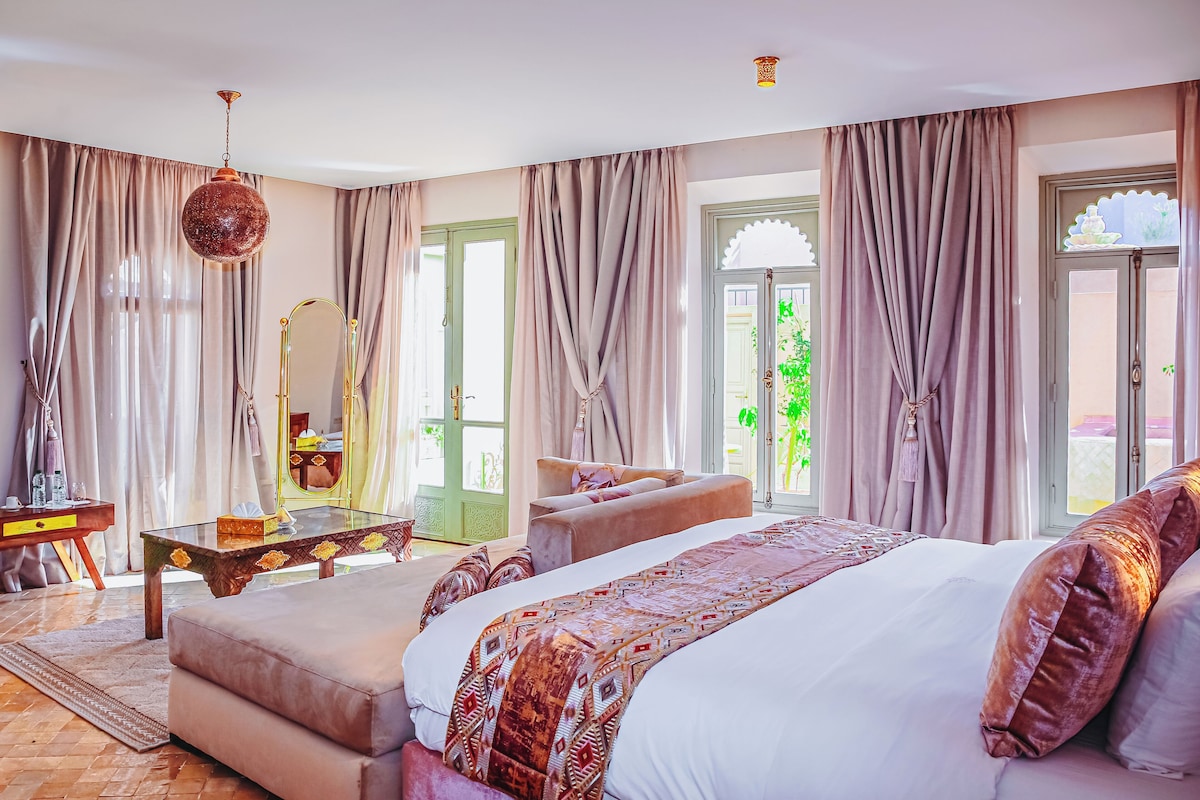 A bedroom with a large bed, a sitting area, a vanity with a mirror, and a view of greenery outside.