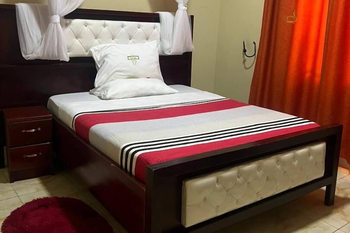 A bedroom with a bed, a nightstand, a red rug, and a curtain.