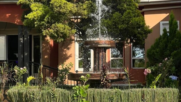 Ein Brunnen, umgeben von gepflegten Hecken und einem großen Baum, mit einem Gebäude mit rotem Dach im Hintergrund.