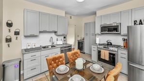 A modern kitchen with grey cabinets, stainless steel appliances, and a dining area with a glass table and cushioned chairs. - Comfort Meets Convenience – 3BR + Free Parking (Boston)