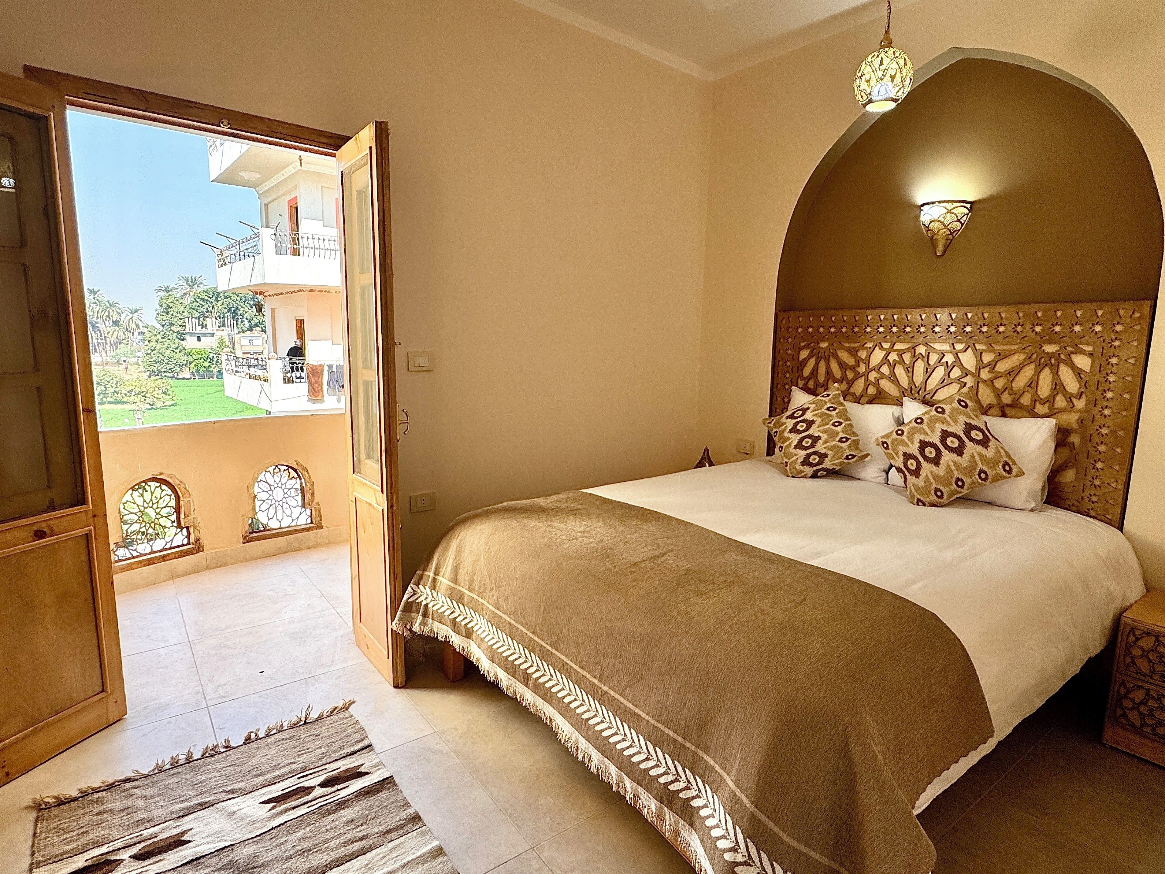 Une chambre avec un grand lit, une vue sur un balcon extérieur accessible par une porte ouverte, et un luminaire décoratif au plafond.