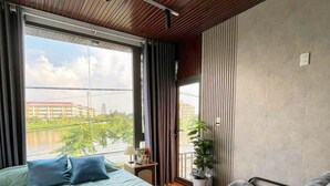 A bedroom with a bed, a chair, a window, and a view of greenery and buildings. - RUBY HOTEL FRONT OF BEACH 2 (Tien Thanh)