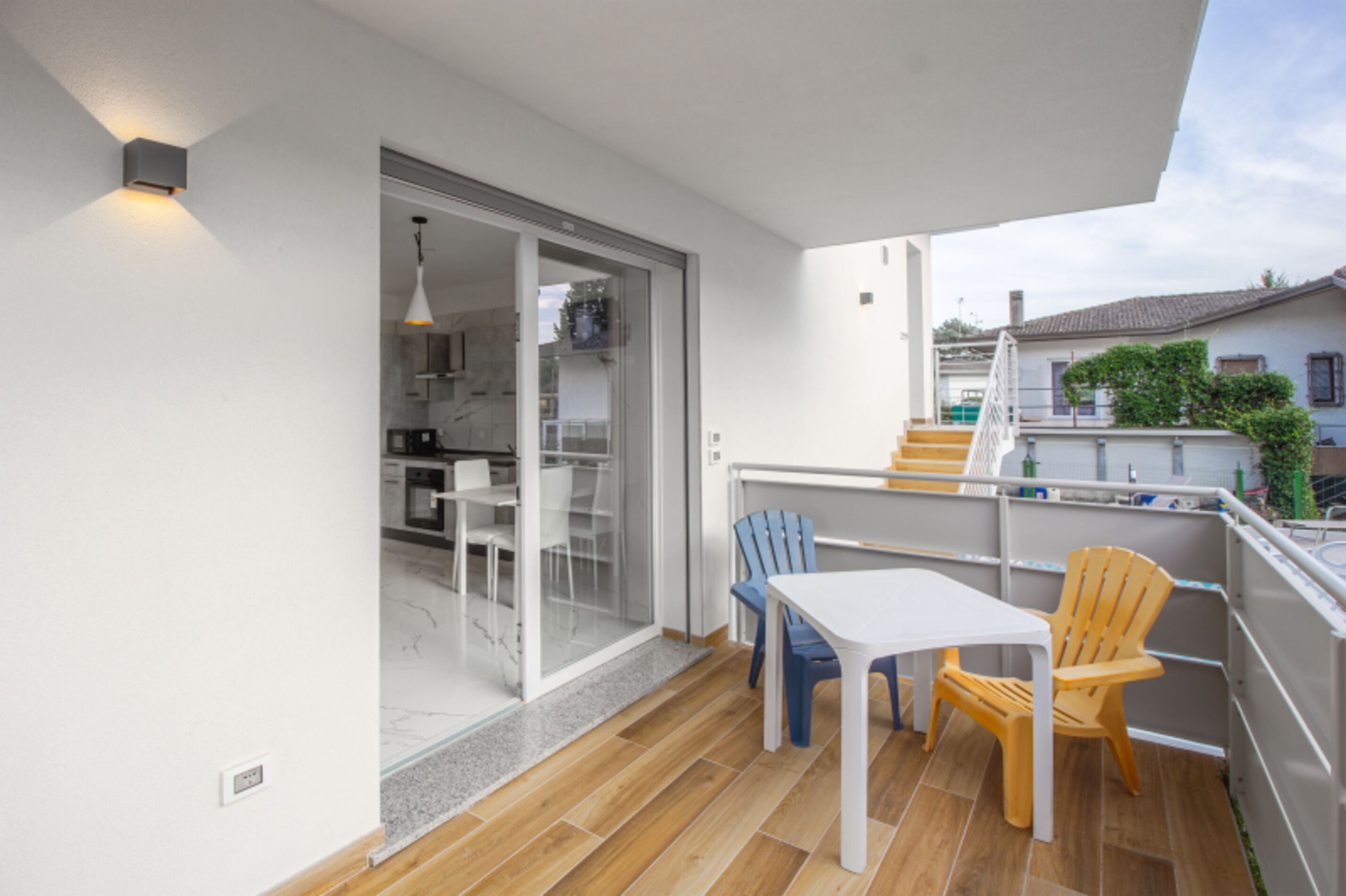 Un balcon avec une table et des chaises, une porte-fenêtre coulissante donnant sur une cuisine et une vue sur les maisons voisines.
