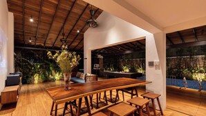 Una habitación amplia con una larga mesa de madera y bancos, un ventilador de techo y vistas a una zona de piscina con plantas.