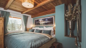 Um quarto aconchegante com teto de madeira, uma cama grande com roupa de cama azul, uma pintura de paisagem emoldurada e um vaso decorativo na parede.