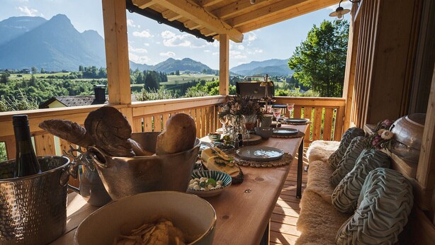 Um deck de madeira com uma mesa posta para refeições, com vista para uma paisagem montanhosa.