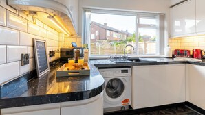 A modern kitchen with a built-in washing machine, a breakfast bar with croissants and orange juice, and a view of the neighborhood through a large window. - Headingley Lodge - Modern Home Near City Centre (Leeds)