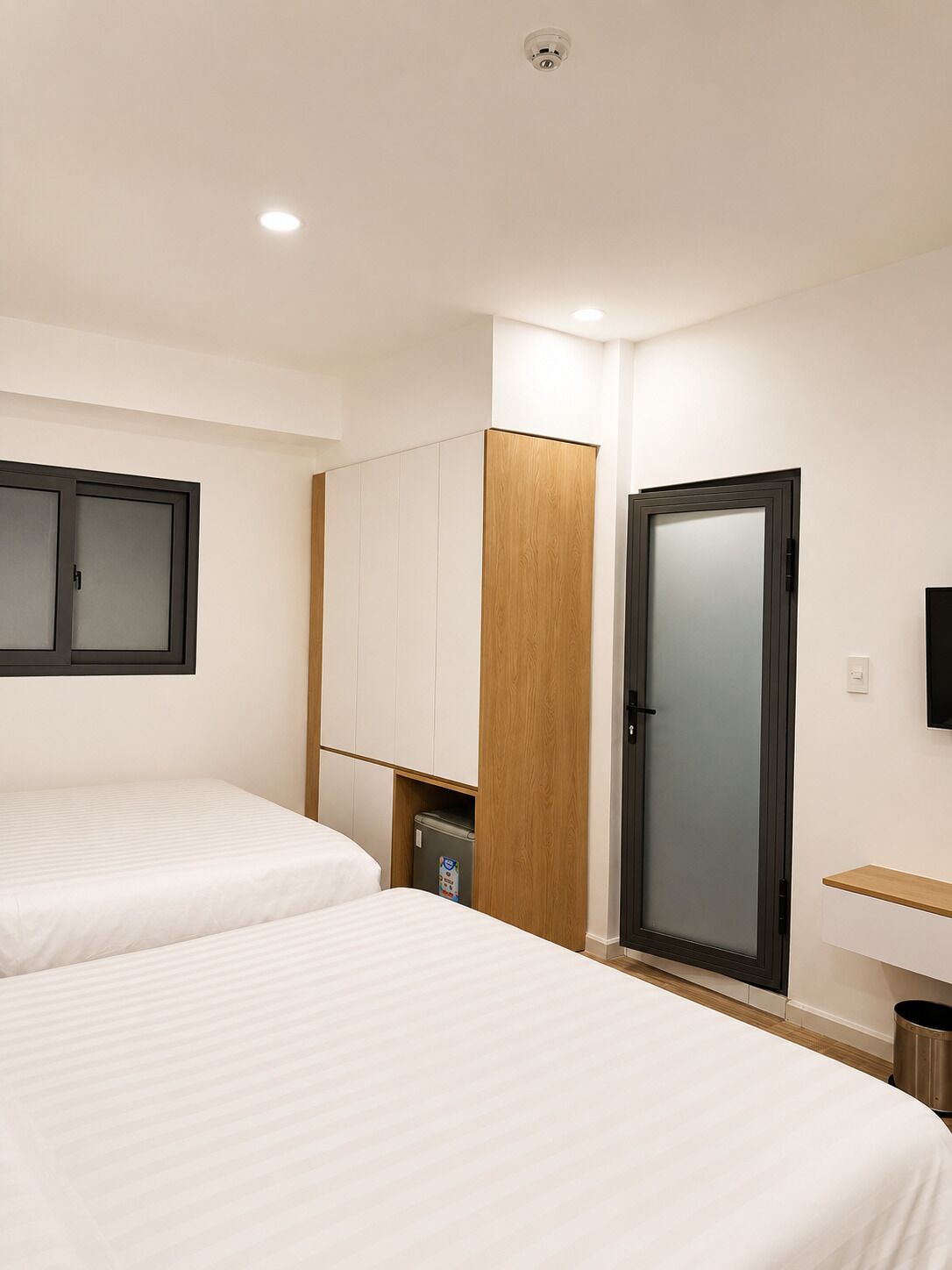 A hotel room with two beds, a wardrobe, a trash bin, and a television on the wall.