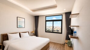 Una camera d'albergo con un letto, un comodino, una lampada, una finestra vista città e uno scaffale con un bollitore.