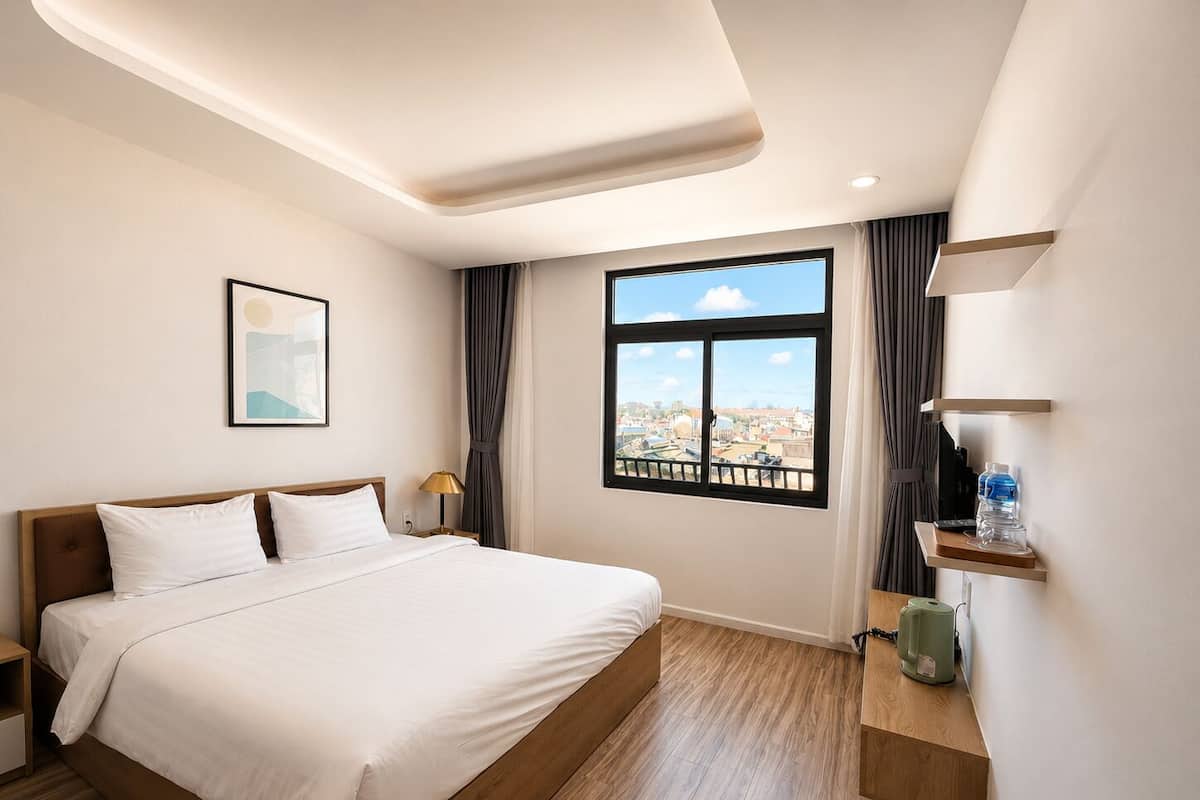 A hotel room with a bed, a nightstand, a lamp, a window with a city view, and a shelf with a kettle.