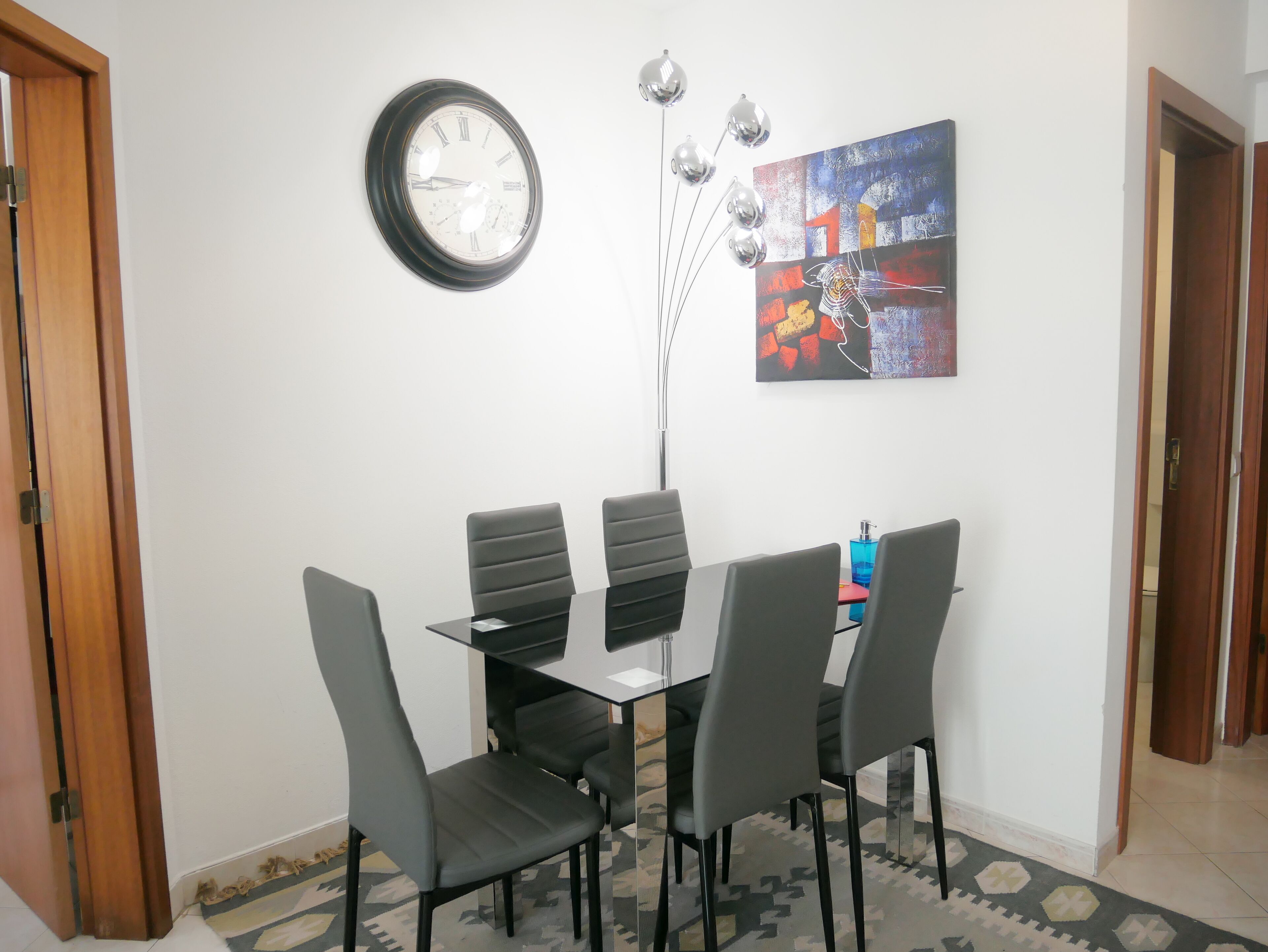 A dining area with a round wall clock, a modern abstract painting, and a glass table with four chairs.