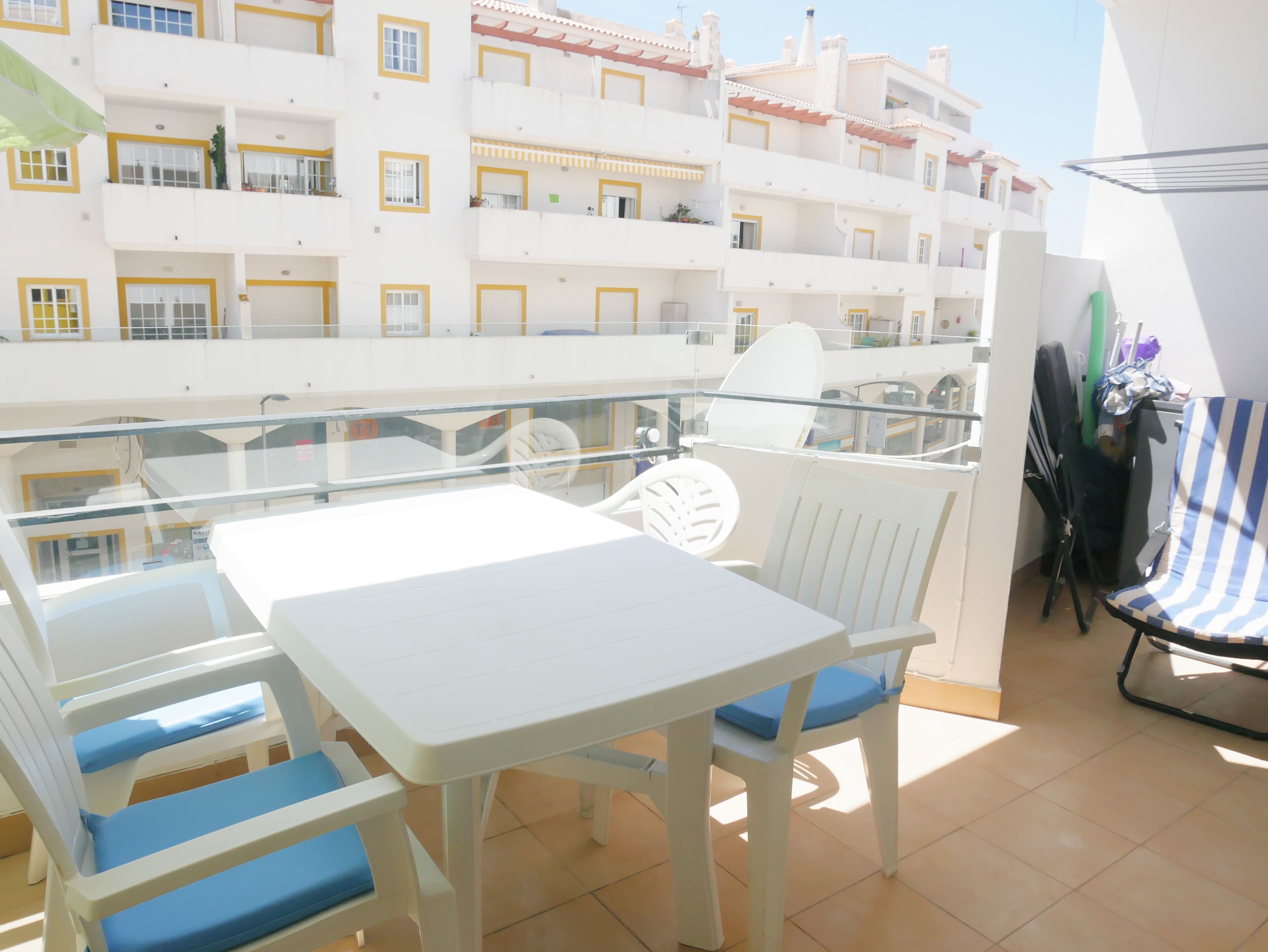 A balcony with a white table and chairs, a glass railing, and a view of multi-story buildings.