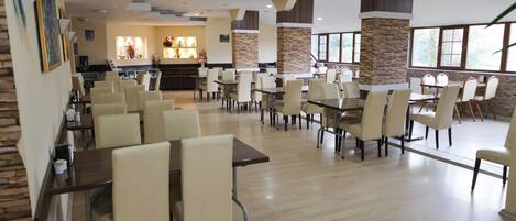 A spacious dining area with multiple tables and chairs, a kitchen in the background, and large windows providing natural light.
