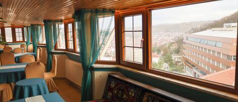 A room with a wooden ceiling, large windows with curtains, a patterned bench, and tables with blue tablecloths.