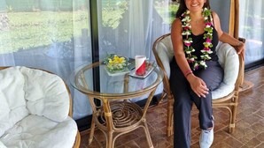 Une personne est assise sur un fauteuil en osier dans une pièce où se trouvent une table en verre, un fauteuil rembourré et une vue sur la verdure à l’extérieur.
