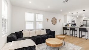 Ein modernes Wohnzimmer mit einer schwarzen Couch, zwei runden Holztischen und einem weißen Teppich. Zwei Fenster mit weißen Jalousien, ein Küchenbereich mit Barhocker und eine Wanduhr sind ebenfalls vorhanden.
