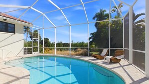 A covered swimming pool with a curved edge, surrounded by a white fence, and equipped with lounge chairs. - Luxury Coastal Retreat | Salt Water Heated Pool (Sanibel)