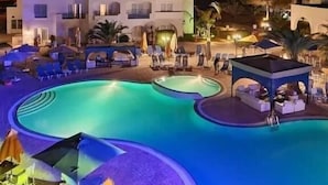 Ein Hotel mit Schwimmbeckenbereich bei Nacht, inklusive Liegestühlen und Sonnenschirmen.