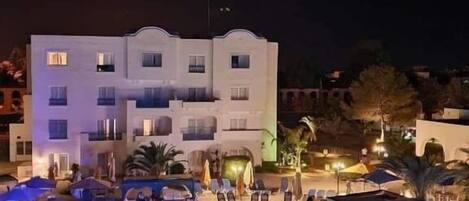 Un hotel con zona de piscina por la noche, con sillones de descanso y sombrillas.