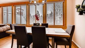 A dining area with a wooden table and chairs, large windows with a view of trees, and a chandelier. - Beautiful Breck Condo, block from Main and Gondola (Breckenridge)