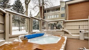 A hot tub in a covered outdoor area with a wooden deck, surrounded by a building with large windows and a view of a snowy landscape. - Beautiful Breck Condo, block from Main and Gondola (Breckenridge)