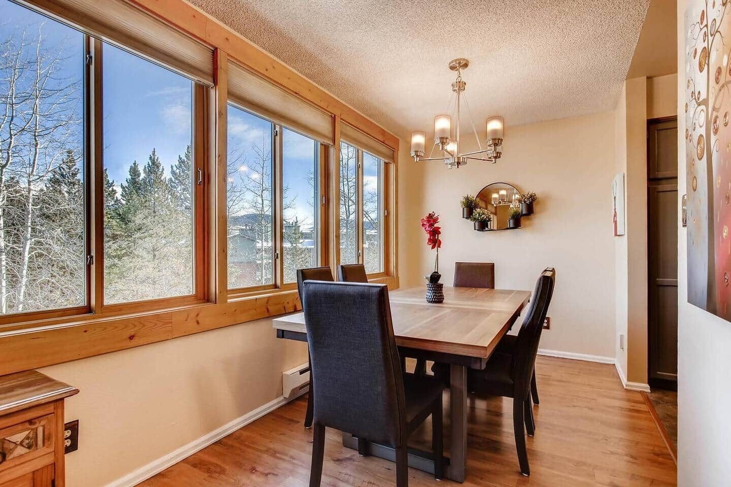 A dining area with a wooden table and chairs, large windows with a view of trees, and a chandelier.