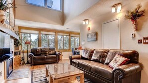A spacious living room with leather sofas, a wooden coffee table, and large windows offering a view of trees. - Beautiful Breck Condo, block from Main and Gondola (Breckenridge)