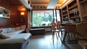 Ein gemütliches Blockhaus-Interieur mit Holzbalkendecke, einem großen Fenster mit Blick auf Bäume, einer Couch, einem Tisch und einem Bücherregal.