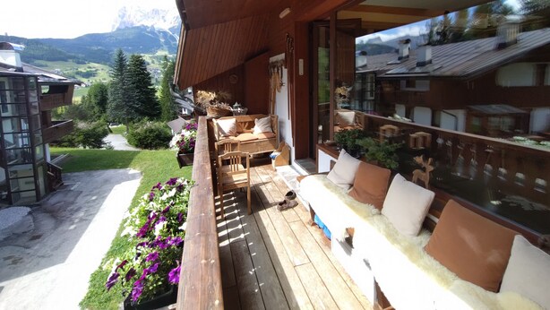 Eine Holzterrasse mit Sitzgelegenheiten im Freien, eine Glaswand mit Blick auf die Berge und ein Garten mit Blumen.