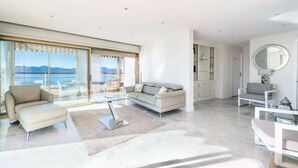 Un salon moderne avec un canapé, un fauteuil et une table basse, de grandes fenêtres offrant une vue sur la mer, et un grand miroir rond accroché au mur.