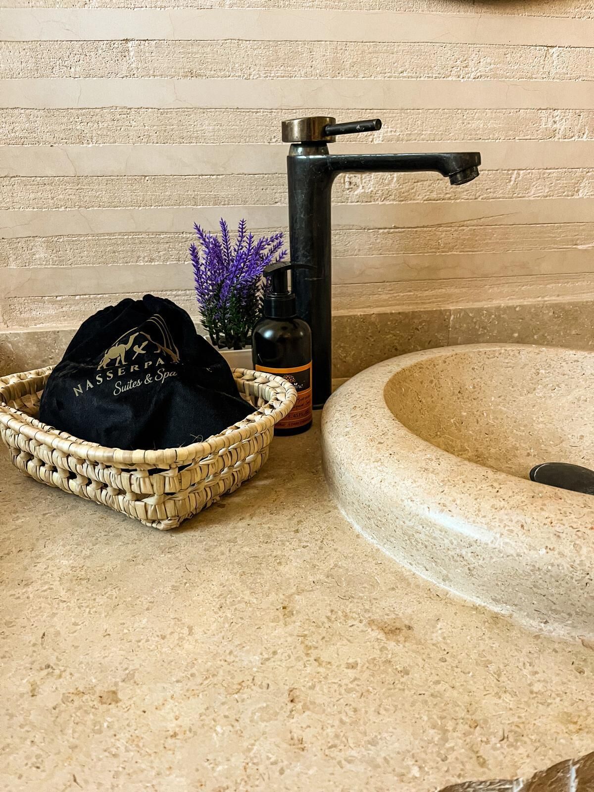 A bathroom with a stone sink, a black faucet, a woven basket with a black bag, and a bottle of soap.