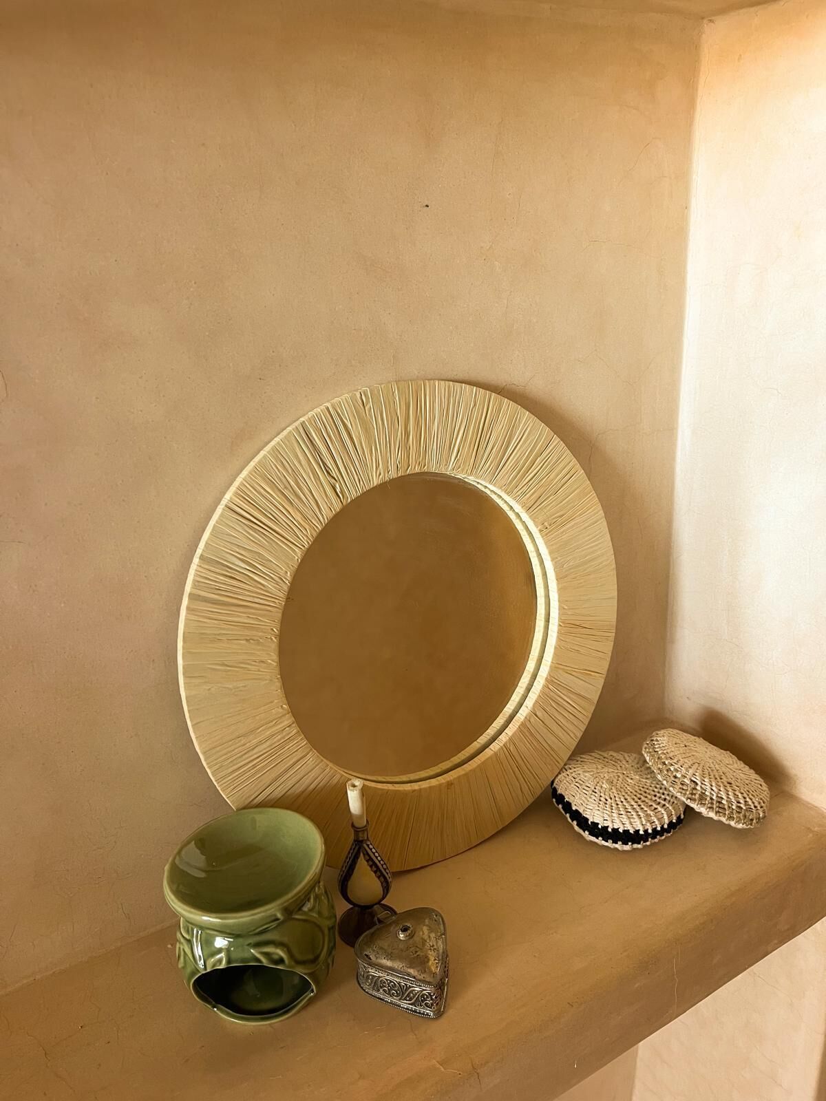 Une étagère avec un miroir rond, une tasse verte, un petit récipient en argent et deux coussins en crochet.