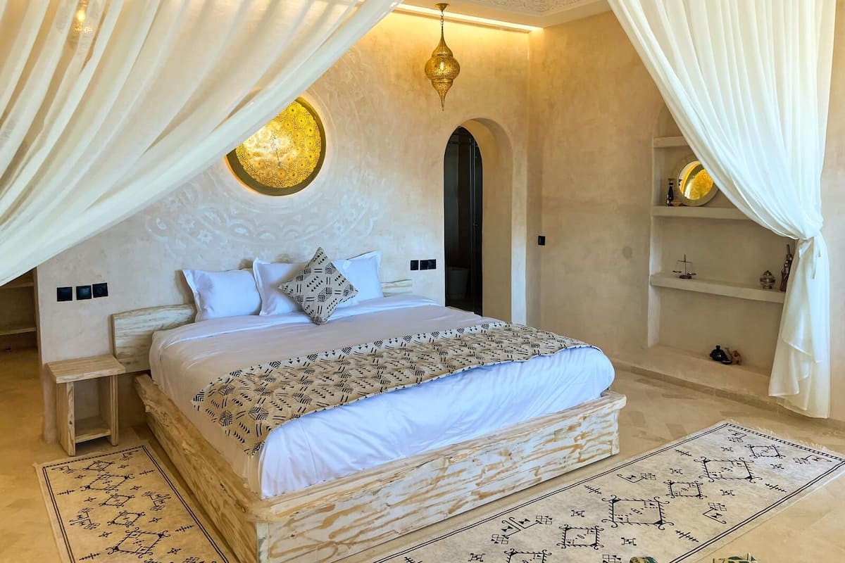 Une chambre à coucher avec un grand lit, un mur décoratif, un lustre et une petite étagère sur laquelle sont posés divers objets.