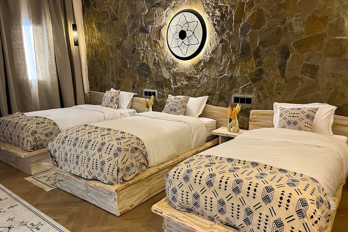 Une chambre d’hôtel avec trois lits, chacun doté de draps à motifs et d’une tête de lit en bois. Un mur en pierre avec un luminaire circulaire est visible derrière les lits.