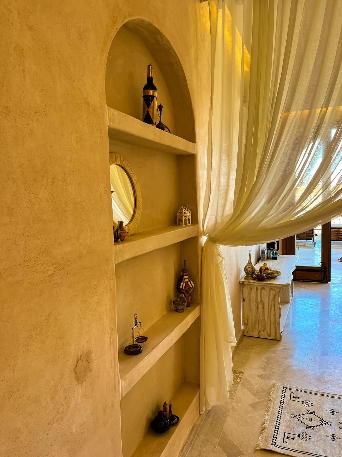 A room with a large arched shelf holding decorative items, a mirror, and a small table with a lamp.