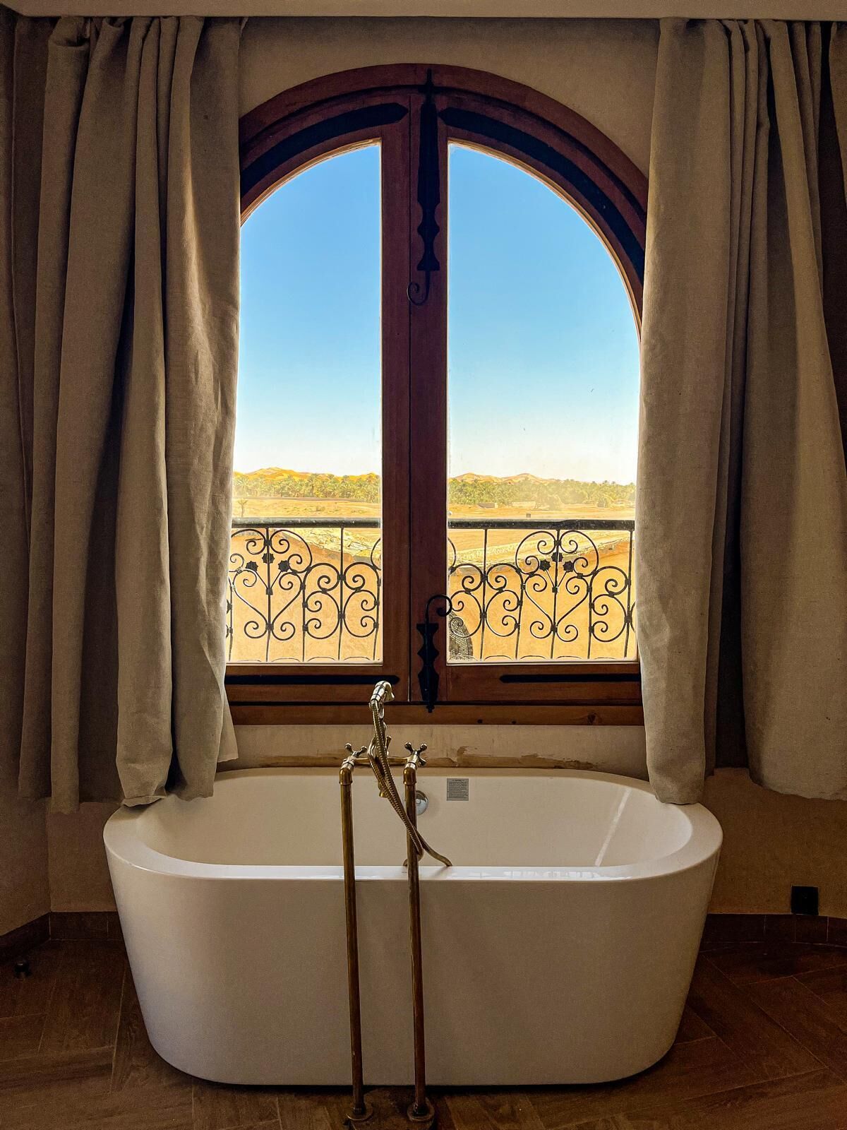 A large freestanding bathtub with a view of a landscape through a window with curtains.