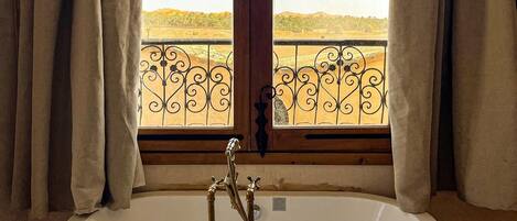A large freestanding bathtub with a view of a landscape through a window with curtains.