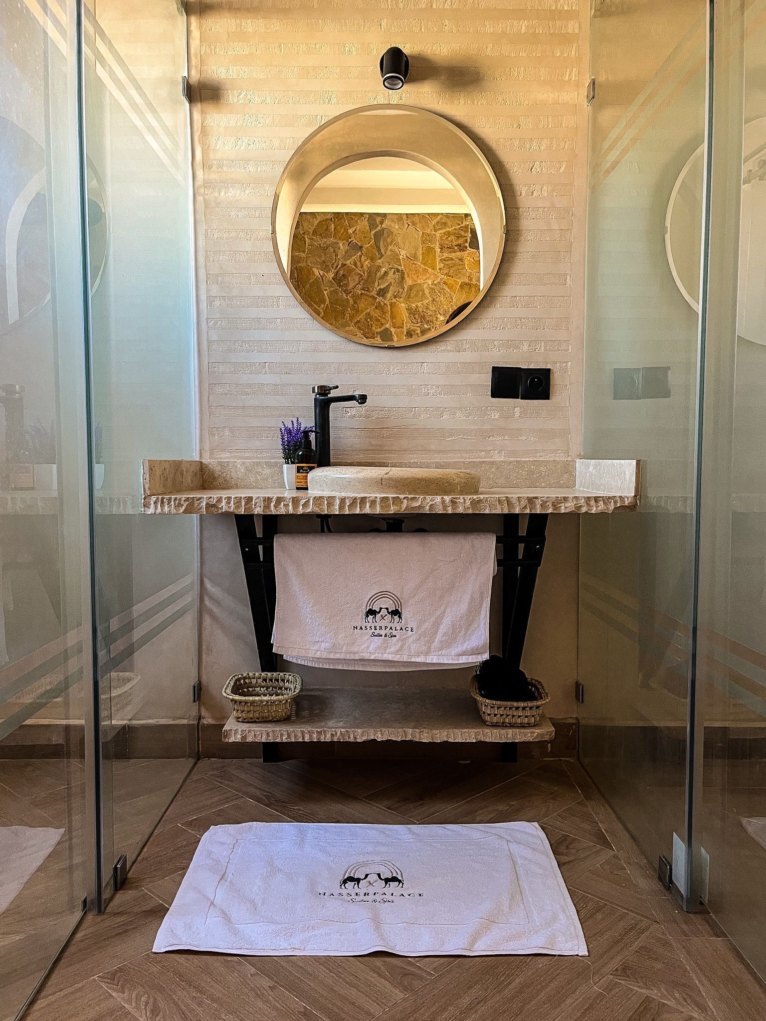 A bathroom with a stone sink, a round mirror, and a towel with a logo on the floor.
