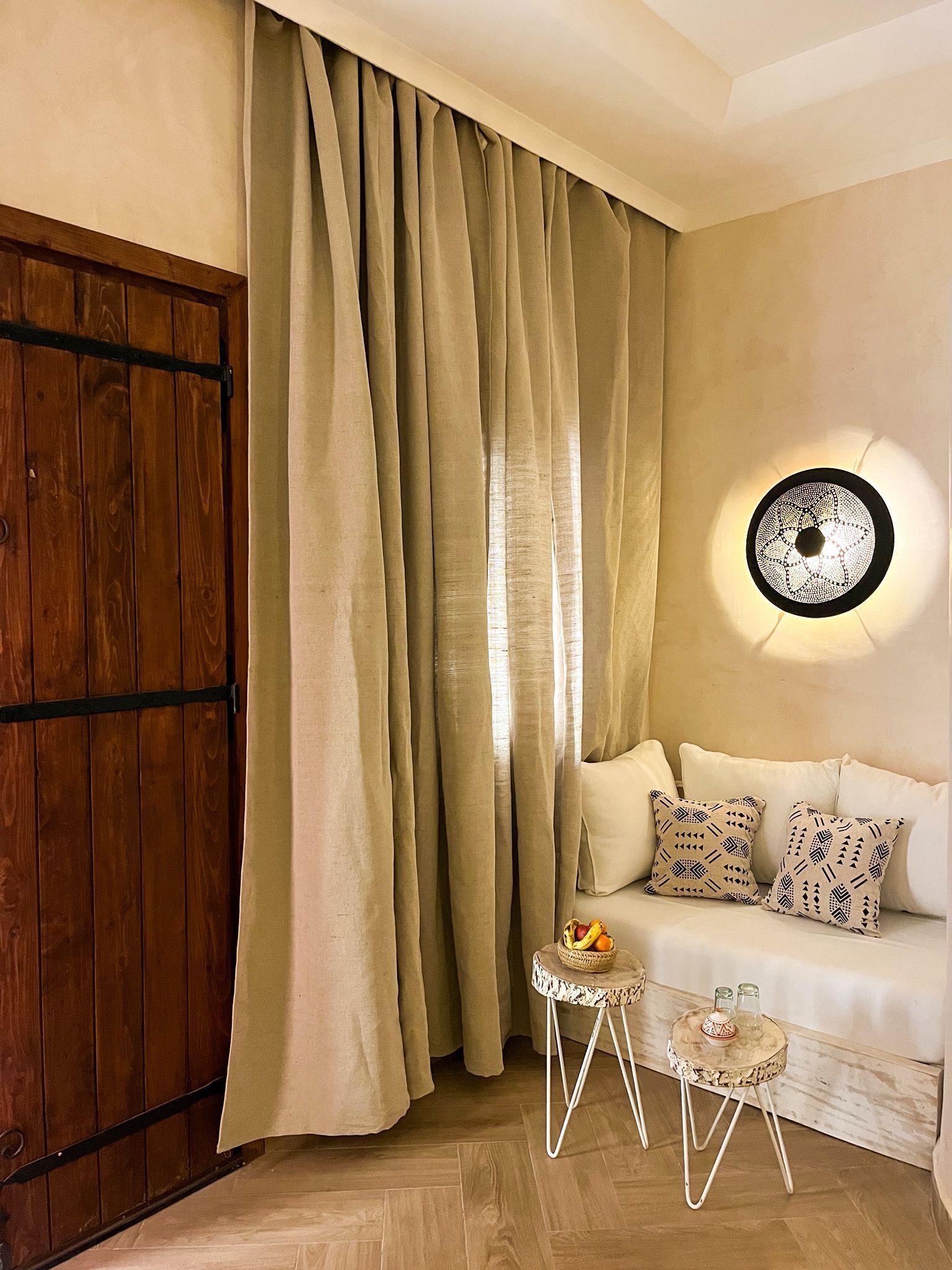 Una habitación con puerta de madera, cortinas color crema, un sofá blanco con cojines estampados, una pequeña mesa redonda con una cesta y vasos, y una lámpara fijada a la pared.